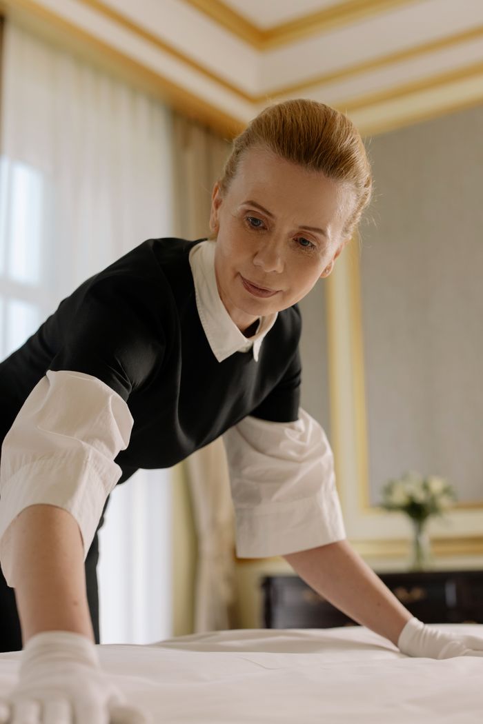 gallery-1 A housekeeper in uniform skillfully arranges bed linen in a luxurious hotel room.