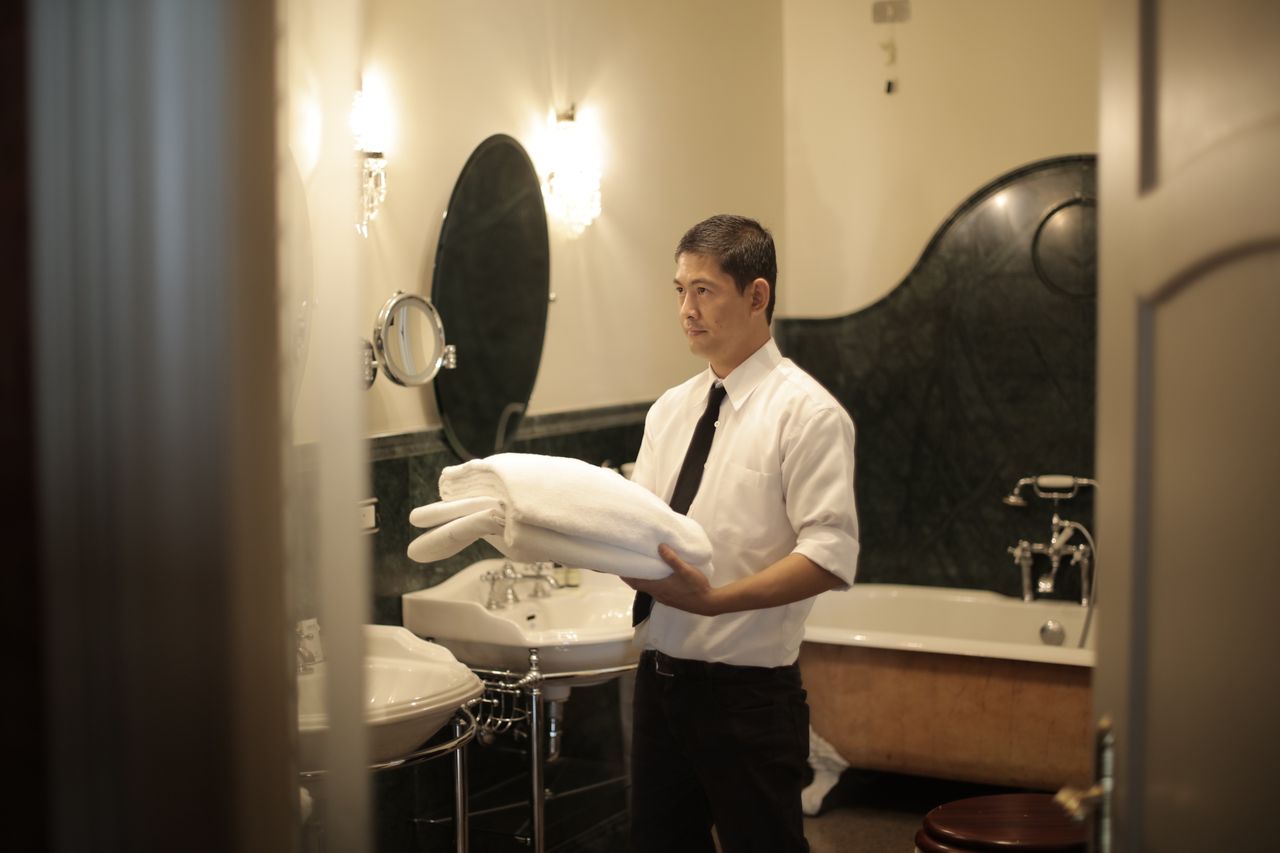 hero-img-02 Side view of young male employee holding stack of white towels while standing in bathroom of luxury hotel